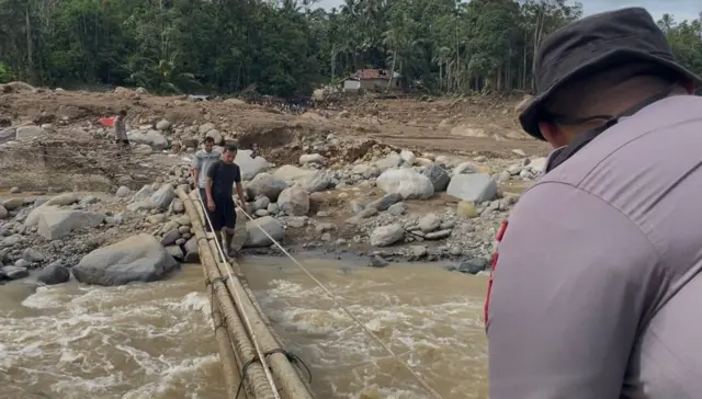 Dua pria sedang menyeberangi jembatan di atas sungai deras setelah banjir bandang Sumbar, dan seorang petugas polisi bersiaga untuk membantu dari seberang.
