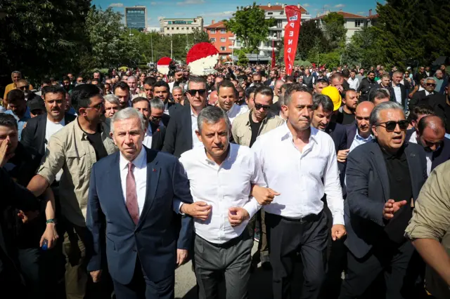 Kol kola girmiş üç erkek, arkada kalabalık, hep birlikte yürüyorlar.