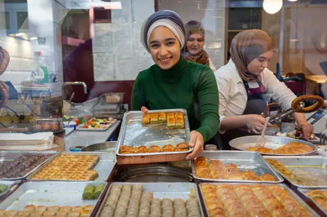 Baklavacı dükkanında çalışan bir genç kadın dikdörtgen bir tepsi baklavayı kameraya doğru uzatıp gülümsüyor. Tepsideki baklavaların çoğu satılmış, azı kalmış. Arkasındaki diğer kadınlar hummalı bir çalışma içinde. 