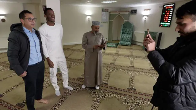 Dango Ouattara, tout de blanc vêtu, pose pour une photo à l'intérieur d'une mosquée, avec l'homme qui prend la photo à droite et un autre homme à l'arrière-plan qui regarde son téléphone.