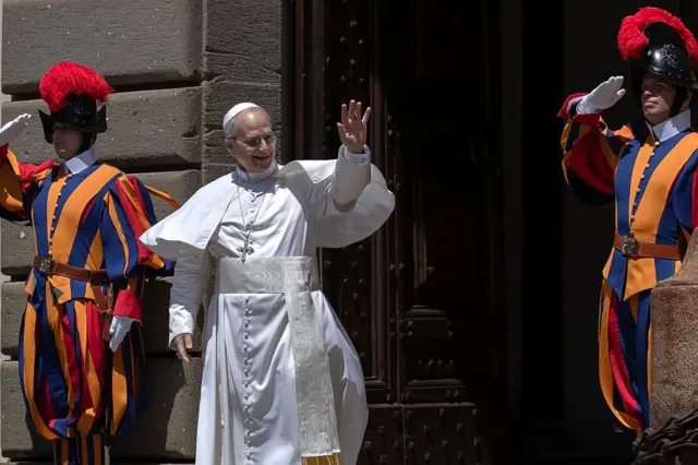 Le Pape Léon XIV lève la main gauche en signe de salutation, deux gardes suisses pontificale se trouvent à ses côtés.