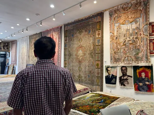 Eric Saddith de espaldas en su tienda alfombras de Teherángeles, con alfombras colgadas de las paredes y tapices con el retrato del último sha de Irán, Mohamed Reza Pahlavi, el 15 de junio de 2026. (Foto: Leire Ventas)
