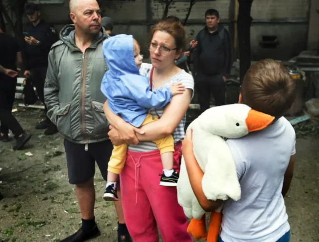 Жінка у рожевих штанях триває на руках дитину у синій куртці. Хлопчик років 9-10 тримає м'яку іграшку-гуся і стоїть спиною до камери. Чоловік у зеленій куртці дивиться у бік. 