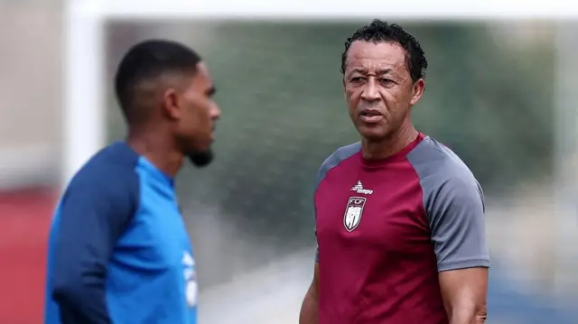 L'entraîneur du Cap-Vert, Bubista, vu de la taille jusqu'au haut du corps pendant une séance d'entraînement, vêtu d'un haut d'entraînement marron à manches courtes grises. Il est net et se tourne vers un joueur flou vêtu de bleu au premier plan