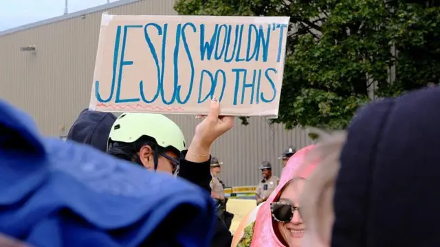 كاثوليك يتظاهرون أمام مركز للمهاجرين ويرفعون لافتة مكتوب عليها بخط اليد 