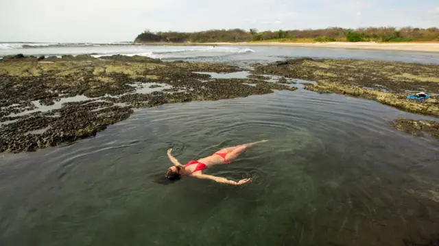 Una mujer con bikini rojo se relaja en una piscina natural. Al fondo se ve la playa.