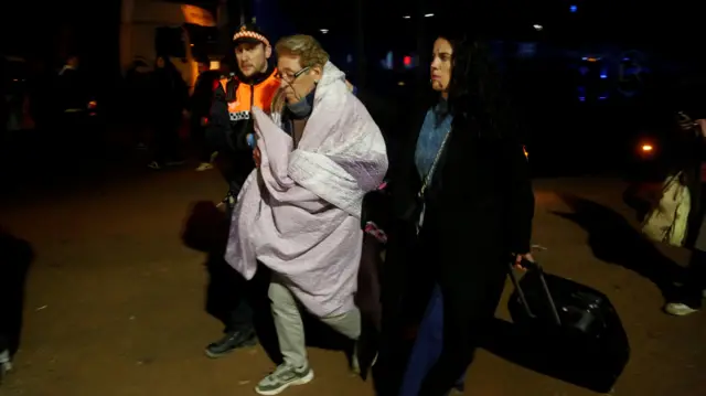 женщина с чемоданом, укрытая пледом, идет в сопровождении человека в форме и девушки