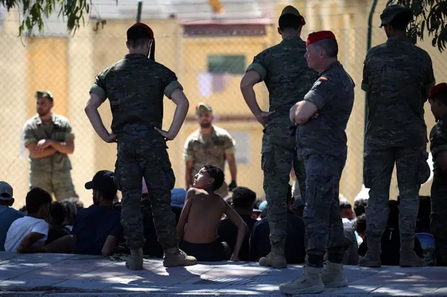 Un grupo de personas sentadas en el suelo al aire libre. A su alrededor un grupo militares españoles de pie los vigila. Entre las personas sentadas, un niño se gira y eleva la mirada hacia uno de los militares. 
