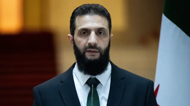 صورة لرجل ذي شعر كثيف داكن اللون ولحية كثيفة وشارب كلاهما داكن اللون أيضاً. يرتدي الرجل سترة ورابطة عنق داكنتين وقميصاً أبيض.