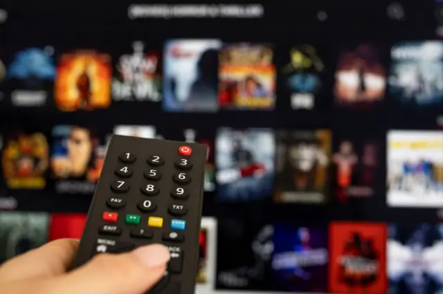 Controle remoto e, atrás, tela de TV com catálogo de filmes 