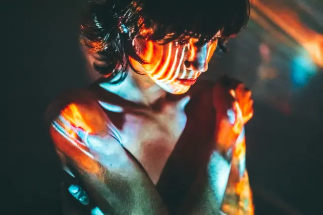 Mujer pensativa en una habitación oscura y con su cuerpo iluminado por imágenes de colores.