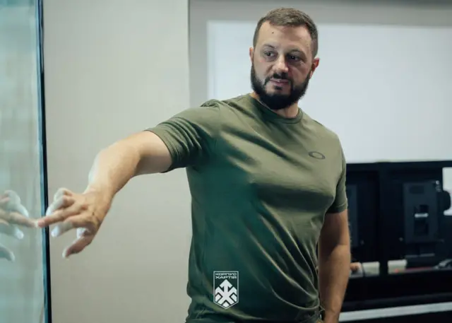 Чоловік в оливковій футболці стоїть у приміщенні біля великого екрана та вказує рукою на її частину. У нижній частині фото видно логотип "Корпус Хартія".