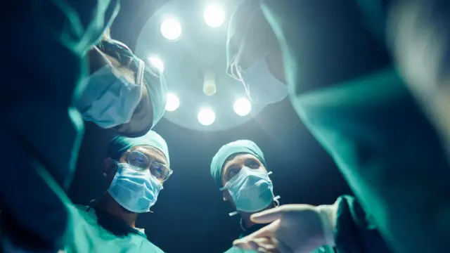 A group of surgeons surround a patient. They are wearing masks and can be seen from the perspective of the patient. 