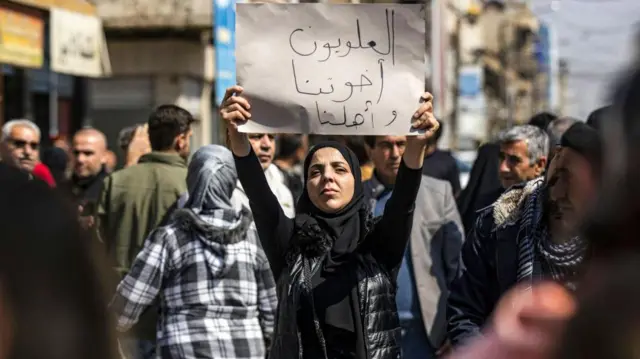 Женщина в окружении других людей стоит на демонстрации и держит плакат, на котором написано по-арабски «Алавиты — наши братья и семья»