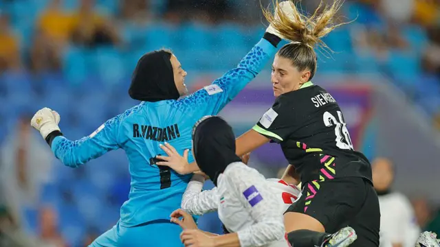 رها یزدانی، دروازه‌بان ایران (سمت چپ)، در جریان بازی ایران و استرالیا در مسابقات جام ملت‌های زنان آسیا ۲۰۲۶ در ورزشگاه گلد کوست، در حالی که تحت فشار رمی سیمسن از استرالیا (سمت راست) قرار دارد، با مشت توپ را از روی ضربه کرنر دور می‌کند.