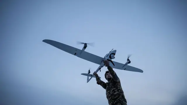 Ukrayna'da havada uçan bir İHA'nın tekerleklerinden tutan bir asker yerde poz veriyor
