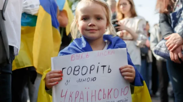 Маленька дівчинка, огорнувшись прапором України, тримає плакат "Говоріть зі мною українською".