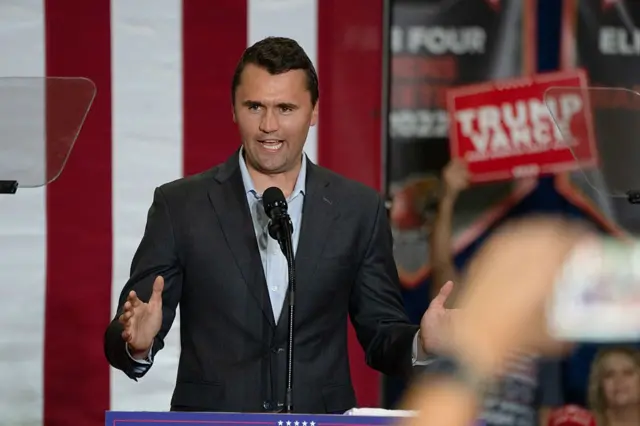 Kirk falando em um púlpito com bandeiras dos Estados Unidos e cartazes escritos "Trump e Vance" no fundo