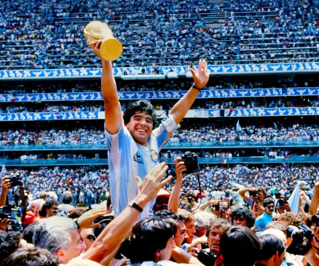 Diego Maradona brandit le trophée de la Coupe du Monde de la FIFA, célébrant sa victoire sous les acclamations des supporters et de ses coéquipiers après la finale de la Coupe du Monde de la FIFA 1986 au Mexique, opposant l'Argentine à l'Allemagne de l'Ouest le 29 juin 1986 au stade Azteca de Mexico.