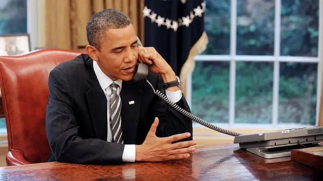باراک اوباما در دفتر بیضی کاخ سفید پشت میز در حال مکالمه با تلفن رومیزی با کت سیاه و کراوات راه‌راه