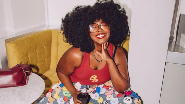 A woman wearing her natural afro hair is smiling at the camera. She's sitting on a yellow corner armchair, slightly leaned forward. She is wearing funky 70s inspired tinted glasses, a red top, and denim trousers with lots of little animals and fruits on!