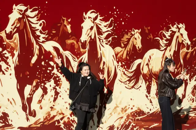 Две женщины позитируют возле мурала с лошадьми. Расцветка мурала красно-желто-белая, напоминающая оттенки огня.