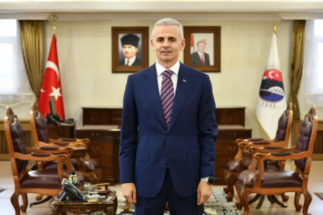 Çiçekli lacivert takım elbiseyle odasında ayakta duruyor.