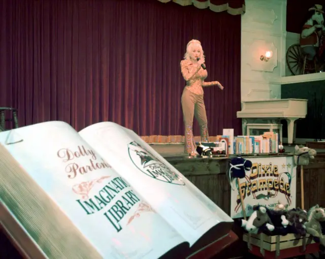 Dolly Parton habla en un escenario al fondo, mientras que en primer plano un gran libro con las palabras "Dolly Parton, Biblioteca Imaginaria" está abierto.