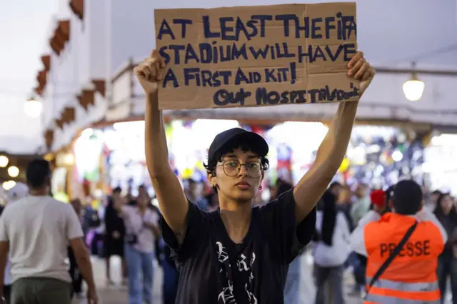Una persona sujeta un cartel en inglés en el que dice que "al menos el estadio de la FIFA tendrá un kit de primeros auxilios".