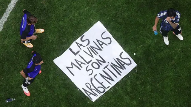Una pancarta que dice "Las Malvinas Son Argentinas" está en el suelo de un campo de juego con tres jugadores de pie a su alrededor.