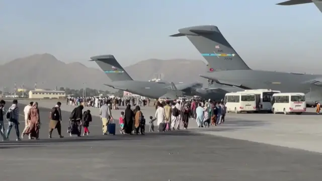 People carrying luggage walk across an airport tarmac towards large US military transport aircraft during an evacuation