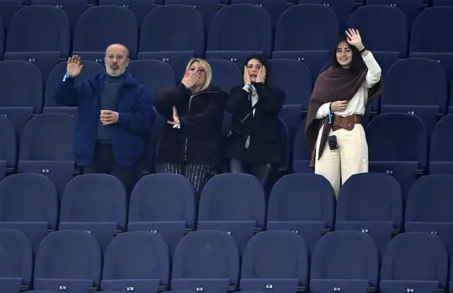 Lacivert koltuklarda bir erkek ve üç kadın yan yana. Heyecanla sahayı izliyorlar.
