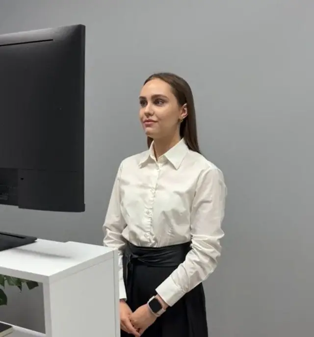 Ведуча онлайн-церемонії Богдана Олех у білій сорочі і чорній спідниці стоїть перед монітором 