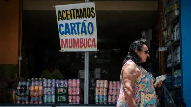 Mulher passacaça níquel pixfrente a comércio com placa dizendo "Aceitamos cartão Mumbuca", moeda social do programacaça níquel pixrenda básica do municípiocaça níquel pixMaricá (RJ)