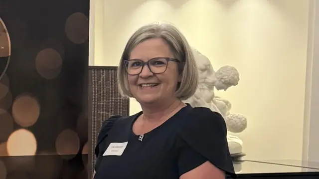 Lisa Ackerman, vêtue d'un élégant haut noir, sourit devant un scuplteur classique.