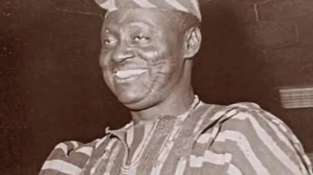 Ayoola Adeleke wọ asọ oke ẹtu ati fila rẹ, o n rẹrin