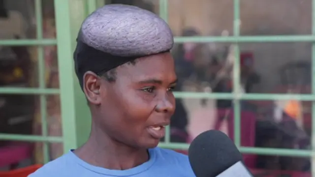 One head and shoulders image of Theresa Zulu wearing blue T-shirt. She dey turn to di right side of di picture and dey tok into microphone wey dey for di bottom right of di frame.