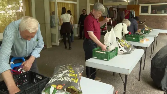 W banku żywności ludzie wybierają produkty z zielonych plastikowych pudełek na stołach i pakują je do toreb na zakupy. W tle widać innych ludzi czekających na swoją kolej.