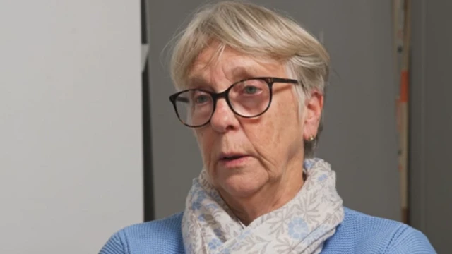 Linda King da una entrevista en video desde su casa. Tiene el pelo corto y rubio, gafas y lleva una bufanda gris claro sobre una blusa morada.