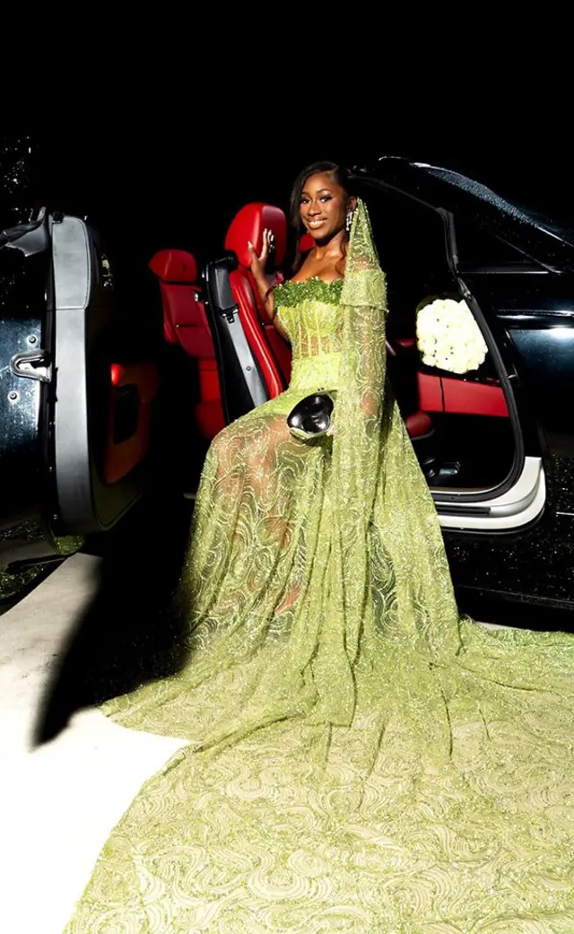 Nian Fisher assise près de la portière ouverte d'une limousine noire. Sa robe verte est ornée d'une traîne disposée devant elle.