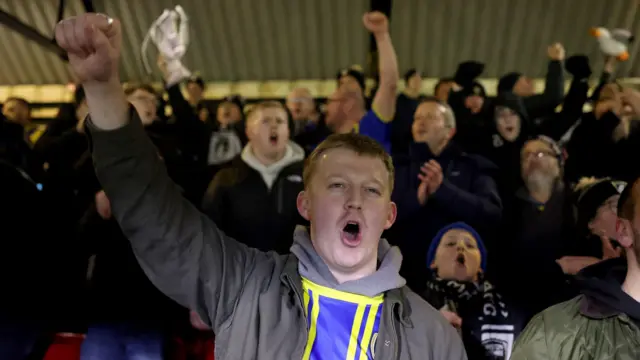 Los aficionados del Weston-super-Mare FC animan a su equipo desde las gradas de Blundell Park durante el partido de la FA Cup contra el Grimsby Town. Delante, un joven con una sudadera gris y una camiseta de rayas amarillas y azules del Weston. Algunos aficionados sostienen versiones pequeñas de la FA Cup.