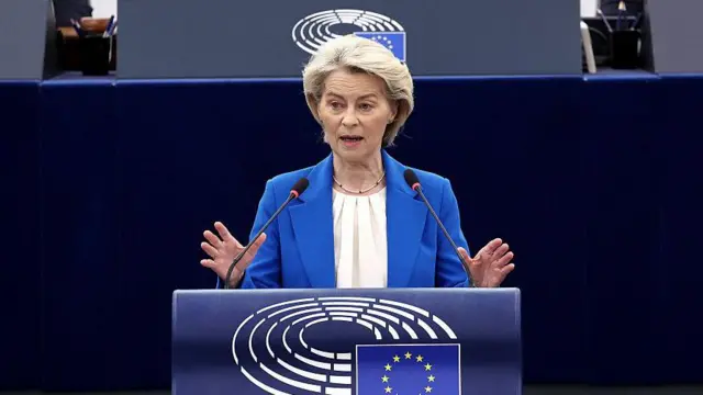 Mavi takım elbiseli sarışın bir kadın AB bayrağı olan bir kürsüde konuşuyor.