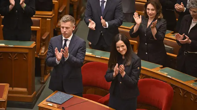 Зала парламенту Угорщини з дерев’яними лавами і столами: у центрі стоять прем’єр Петер Мадяр разом із Анітою Орбан, міністеркою закордонних справ і торгівлі; вони також плескають у долоні. Позаду стоять інші учасники, які теж аплодують.