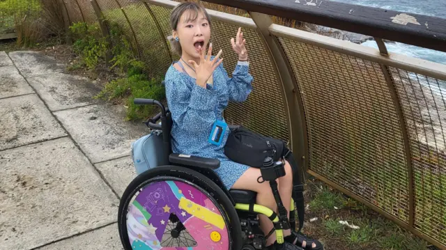 یوه کوریایۍ ښځه په ویلچېیر کې په شنو جامو کې ناسته ده. ټایرونه یې رنګ شوي او د کامرې پر لور یې خوله وازه نیولې ده.