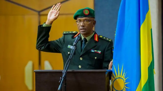 Innocent Kabandana yambaye ikositimu ya gisirikare y'ibara ry'icyatsi kibisi, ishati, karuvati n'ingofero y'icyatsi kibisi ihengetse. Ukuboko kumwe gufashe ku ibendera ry'u Rwanda, mu gihe ikiganza cy'ukundi kuboko kizamuye. Ahagaze imbere y'indangururamajwi n'ameza maremare yo kuvugiraho ijambo. Inyuma ye hari urukuta rw'inzu. Ifoto yo mu mwaka wa 2019.