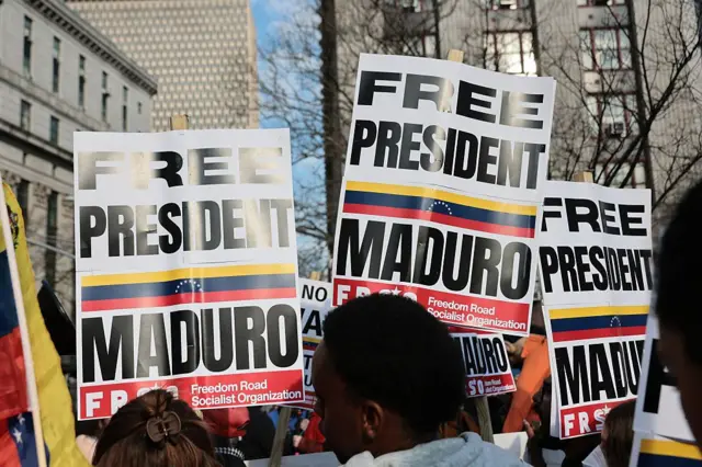 Plano cerrado de personas de espaldas manifestándose. Llevan pancartas. Se ven tres de ellas. En todas se lee lo mismo: "Free President Maduro", "Liberen al presidente Maduro". A lo largo de la pancarta también se ve la bandera venezolana.