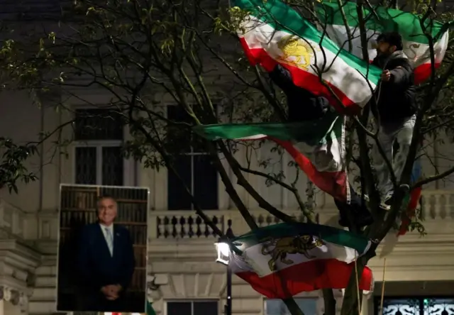 夜のロンドン。白い壁の建物の前、緑?白?赤に金色で獅子と太陽が描かれた旗を、木に登った男性が広げている。スーツ姿に笑顔のパーレヴィー氏の大きい写真も掲げられている