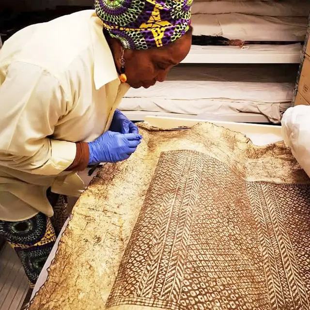 Mulenga Kapwepwe, du Women's History Museum of Zambia, vêtue d'un bandeau vert, violet et jaune à motifs africains, d'une chemise à manches longues crème et de gants en latex bleus, se penche dans un musée suédois pour examiner les motifs complexes des manteaux Batwe.