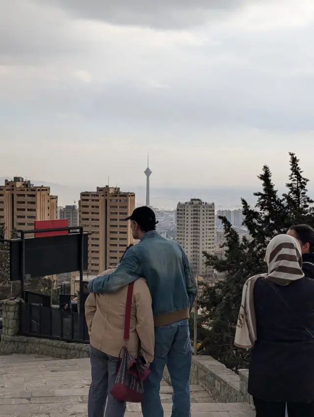 یک مرد دستش را روی شانه زنی گذاشته در حالی که در پس‌زمینه برج میلاد و دود در آسمان تهران دیده می‌شود