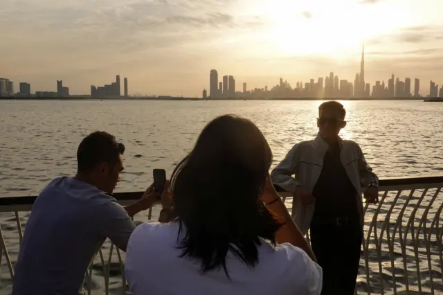 Trzy osoby robią zdjęcia zachodu słońca w Dubaju. Za nimi widać wody zatoki, a na dalszym tle rząd wieżowców - w tym Burdż Chalifę, najwyższy budynek świata. 